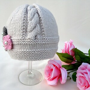 Puede incluir: Un gorro de punto gris claro con un patr&oacute;n de trenza y un adorno de flor rosa. El gorro se muestra en un soporte de vidrio transparente.
