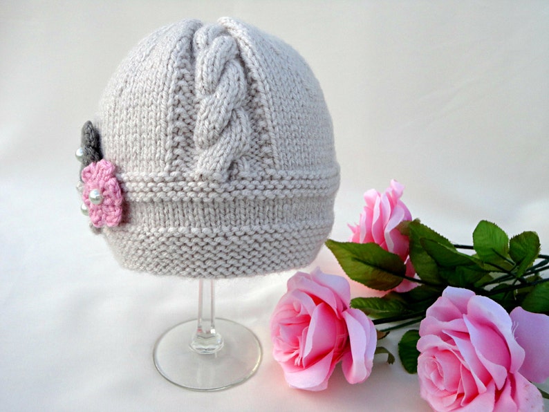 Puede incluir: Un gorro de punto gris claro con un patr&oacute;n de trenza y un adorno de flor rosa. El gorro se muestra en un soporte de vidrio transparente.