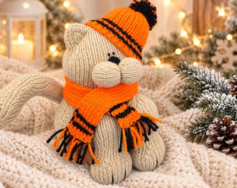 Op de afbeelding: Een beige gebreide kattenknuffel met een oranje en zwart gestreepte muts met een pompon en een bijpassende oranje en zwarte sjaal met franjes. De kat heeft kleine zwarte ogen en een zwarte neus, met een getextureerd gebreid lichaam.
