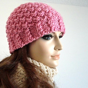 May include: A pink knitted beanie hat with a textured pattern. The hat is worn by a person with long brown hair.