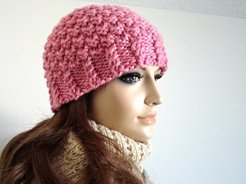 May include: A pink knitted beanie hat with a textured pattern. The hat is worn by a person with long brown hair.
