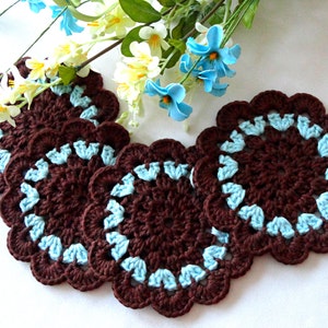 May include: Four brown crocheted coasters with a blue trim. The coasters are round and have a scalloped edge.