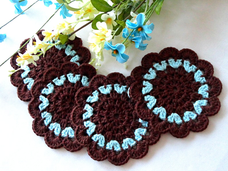 May include: Four brown crocheted coasters with a blue trim. The coasters are round and have a scalloped edge.