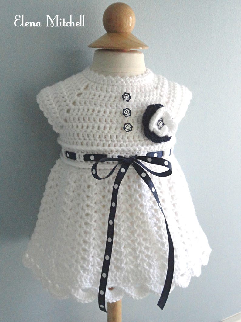 May include: White crocheted baby dress with a black and white polka dot ribbon tied around the waist. The dress has a scalloped hem and three black buttons down the front.