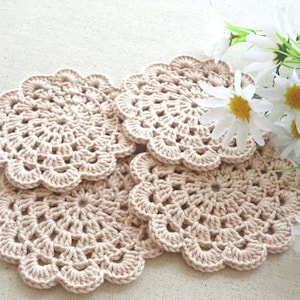 May include: Four crocheted beige coasters with a delicate floral pattern. The coasters are arranged in a stack, with the top coaster slightly offset from the others.