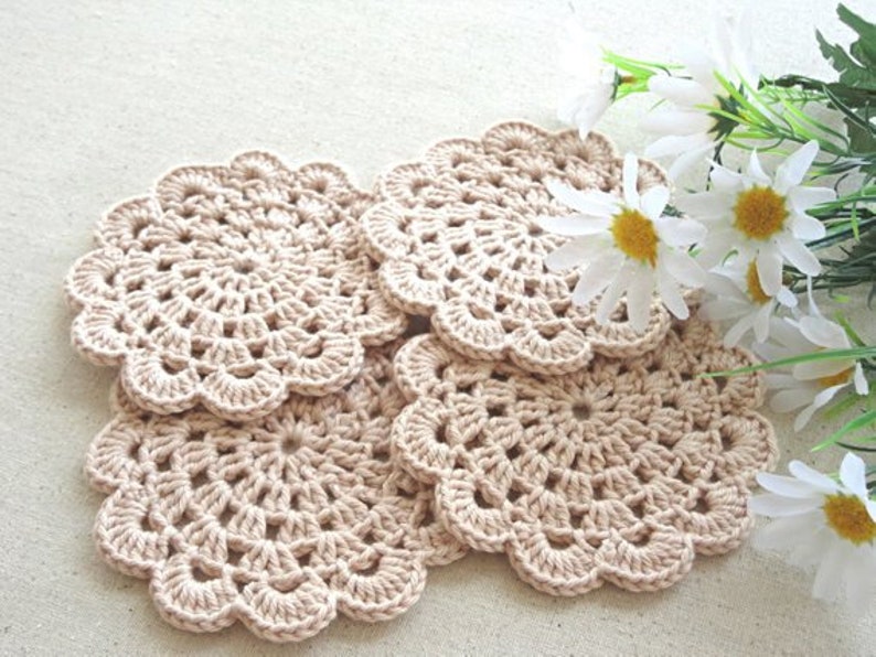 May include: Four crocheted beige coasters with a delicate floral pattern. The coasters are arranged in a stack, with the top coaster slightly offset from the others.