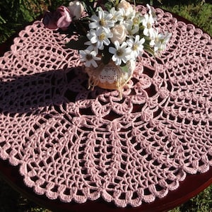 Peut inclure: Un chemin de table en crochet rose d&eacute;licat avec un bord festonn&eacute;. Le chemin de table est pos&eacute; sur une table en bois fonc&eacute; et pr&eacute;sente un motif floral au centre.