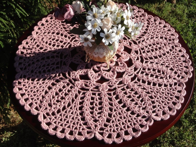 Peut inclure: Un chemin de table en crochet rose d&eacute;licat avec un bord festonn&eacute;. Le chemin de table est pos&eacute; sur une table en bois fonc&eacute; et pr&eacute;sente un motif floral au centre.