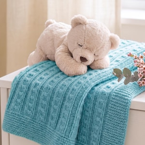 May include: A plush beige teddy bear with closed eyes resting on a folded teal blue knitted blanket. The blanket features a textured cable knit pattern with vertical columns and a wide garter stitch border along the edges.
