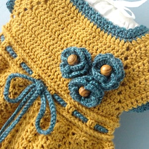 May include: A crocheted baby dress in mustard yellow with a blue crocheted trim and a blue bow. The dress has three blue crocheted flowers with wooden buttons in the center.