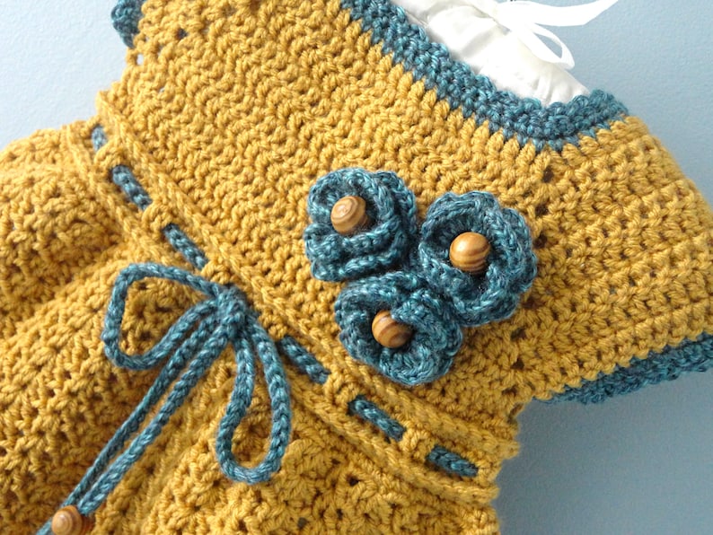 May include: A crocheted baby dress in mustard yellow with a blue crocheted trim and a blue bow. The dress has three blue crocheted flowers with wooden buttons in the center.