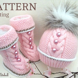 May include: Pink knitted baby hat and booties with a white fur pom pom on the hat. The hat and booties feature a cable knit pattern and pink pearl embellishments.