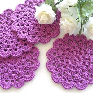 May include: Five purple crocheted coasters with a scalloped edge. The coasters are made with a delicate stitch pattern.