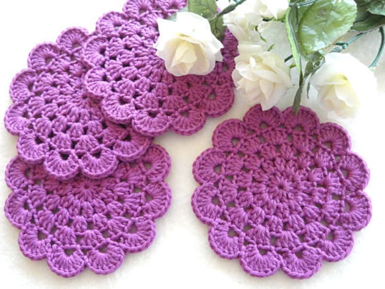 May include: Five purple crocheted coasters with a scalloped edge. The coasters are made with a delicate stitch pattern.