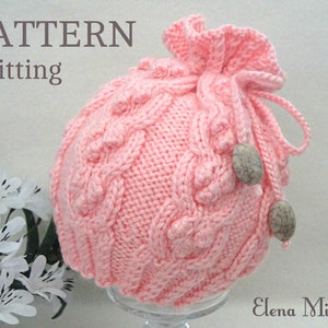 May include: A pink knitted hat with a cable knit pattern and a drawstring closure. The hat has two decorative beads on the side.