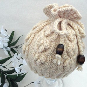 May include: A cream-colored knitted hat with a drawstring and wooden beads. The hat has a cable knit pattern and a ruffled top.