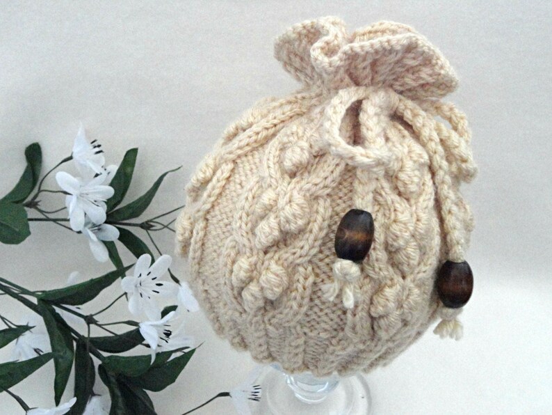 May include: A cream-colored knitted hat with a drawstring and wooden beads. The hat has a cable knit pattern and a ruffled top.