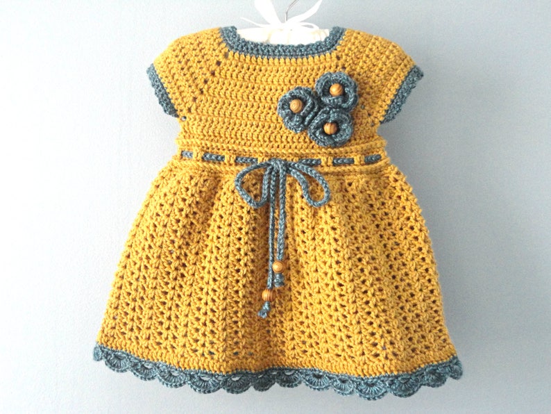 May include: A yellow crocheted dress with a gray crocheted trim and a gray crocheted flower detail. The dress has a tie at the waist.