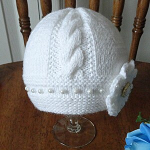 Puede incluir: Sombrero blanco de punto con un patr&oacute;n de trenza y un adorno de flor blanca. El sombrero est&aacute; decorado con una fila de perlas blancas.