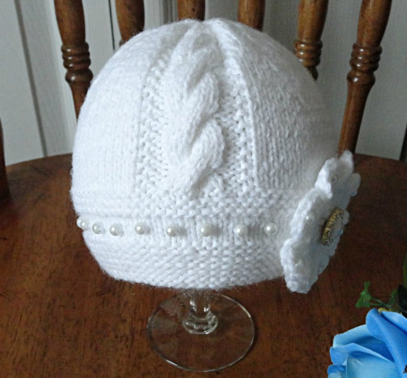 Puede incluir: Sombrero blanco de punto con un patr&oacute;n de trenza y un adorno de flor blanca. El sombrero est&aacute; decorado con una fila de perlas blancas.