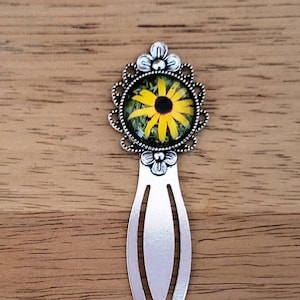 May include: A silver metal bookmark with a decorative top featuring a yellow and black flower design. The flower is set within a circular frame with an ornate, filigree-style border. The bookmark has a simple, elongated shape.