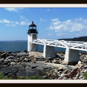 May include: A white lighthouse with a black roof and a white bridge leading to it. The lighthouse is on a rocky island with the ocean in the background.