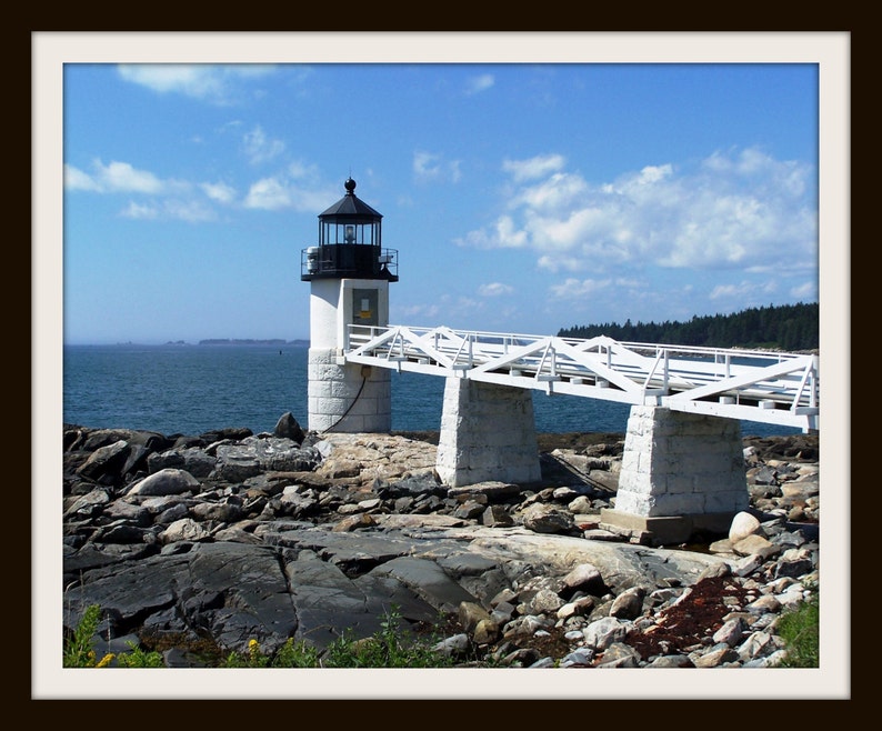May include: A white lighthouse with a black roof and a white bridge leading to it. The lighthouse is on a rocky island with the ocean in the background.