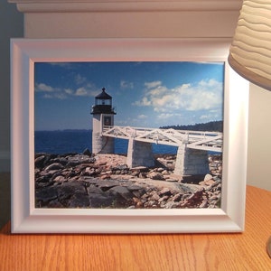 May include: A framed photograph of a white lighthouse with a bridge leading to it. The lighthouse is on a rocky island with the ocean in the background. The sky is blue with white clouds.