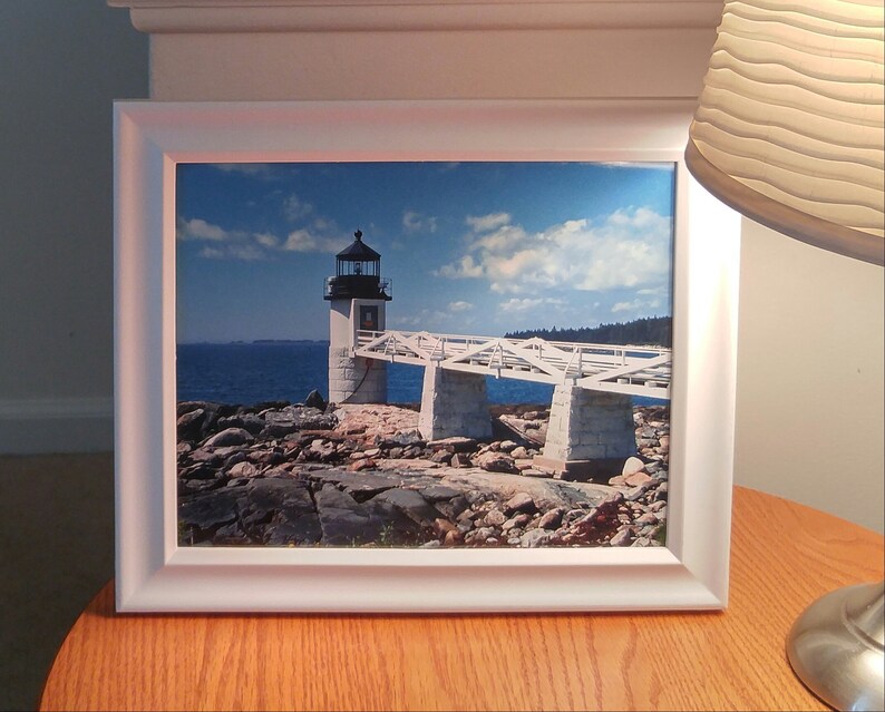 May include: A framed photograph of a white lighthouse with a bridge leading to it. The lighthouse is on a rocky island with the ocean in the background. The sky is blue with white clouds.