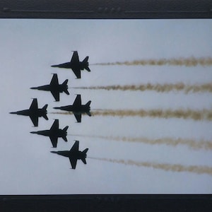 May include: Six black fighter jets flying in formation, leaving white smoke trails against a light blue sky.