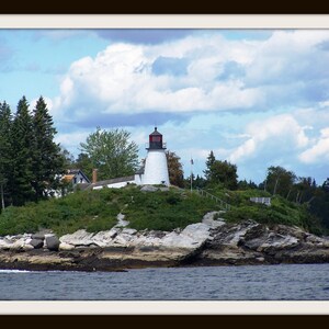 May include: A white lighthouse with a red roof stands on a rocky island. The lighthouse is surrounded by green trees and bushes. The sky is blue with white clouds.