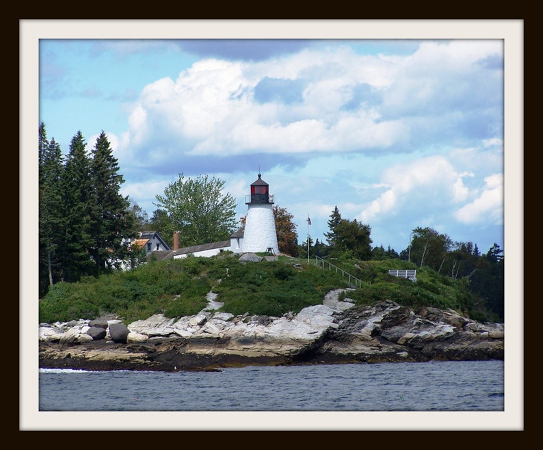 May include: A white lighthouse with a red roof stands on a rocky island. The lighthouse is surrounded by green trees and bushes. The sky is blue with white clouds.