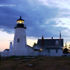 May include: A white lighthouse with a black roof and a light on top. The lighthouse is on a hill overlooking the ocean. There is a white house with a black roof to the right of the lighthouse. The sky is blue and there are clouds in the sky. The text "Mountains to Sea Photos" is in the top left corner of the image.