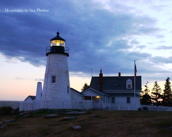 May include: A white lighthouse with a black roof and a light on top. The lighthouse is on a hill overlooking the ocean. There is a white house with a black roof to the right of the lighthouse. The sky is blue and there are clouds in the sky. The text "Mountains to Sea Photos" is in the top left corner of the image.