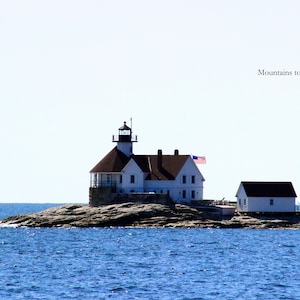 May include: A white lighthouse with a brown roof and an American flag waving in the wind. The lighthouse is on a rocky island surrounded by blue water. The image includes the text "Mountains to Sea Photos".