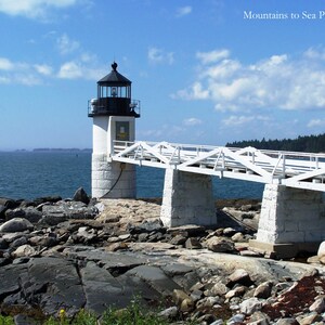 May include: A white lighthouse with a black roof and a white bridge leading to it. The lighthouse is on a rocky shore with the ocean in the background.
