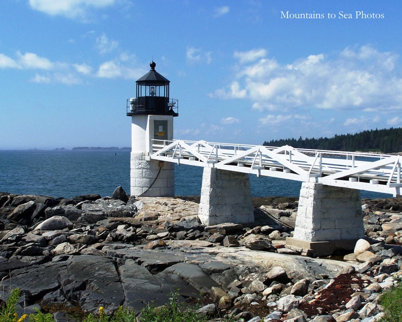 May include: A white lighthouse with a black roof and a white bridge leading to it. The lighthouse is on a rocky shore with the ocean in the background.
