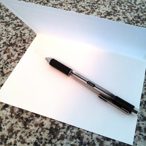 May include: A blank white card with a silver pen on top of it. The card is folded in half and the pen is lying on the inside of the card.