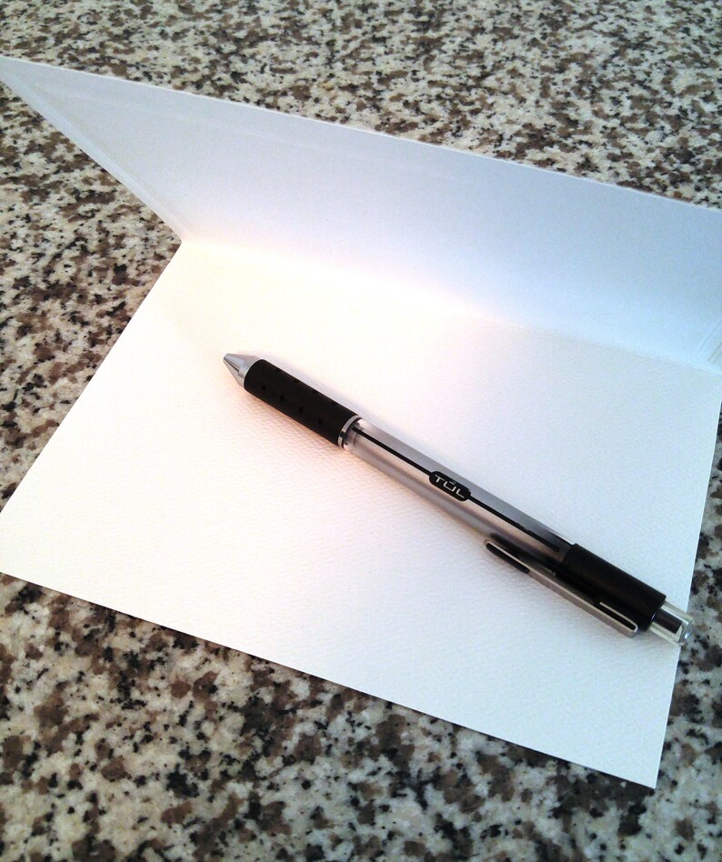 May include: A blank white card with a silver pen on top of it. The card is folded in half and the pen is lying on the inside of the card.