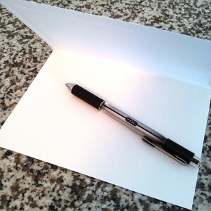May include: A blank white card with a silver pen on top of it. The card is folded in half and the pen is lying on the inside of the card.