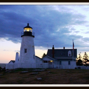 May include: A white lighthouse with a black roof and a light on top. The lighthouse is on a rocky coast with a house and a flagpole in the background. The sky is blue with clouds.