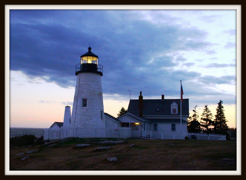 May include: A white lighthouse with a black roof and a light on top. The lighthouse is on a rocky coast with a house and a flagpole in the background. The sky is blue with clouds.