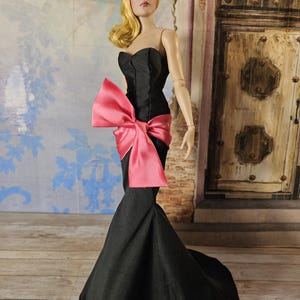 Puede incluir: Un vestido negro sin tirantes con un gran lazo rosa en la cintura. El vestido tiene un corpiño ajustado y una falda fluida.