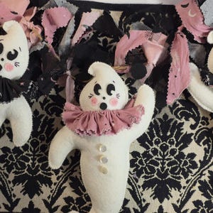 Könnte beinhalten: Drei handgefertigte Geisterpuppen mit weißen Körpern, schwarz-rosa Rüschenkragen und Knopfakzenten. Die Geister haben rosige Wangen und überraschte Ausdrücke. Sie sind vor einem schwarz-weißen Damastmuster und rosa Stoff platziert.