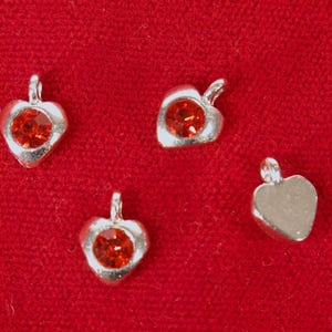 May include: Four silver heart-shaped charms with red rhinestone accents. The charms have a loop at the top for attaching to a necklace or bracelet.