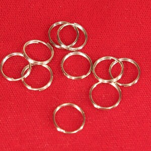 May include: A collection of eight silver-colored metal split rings arranged in a scattered pattern. Each ring features a double-loop coil design, commonly used for keychains or jewelry attachments, with a smooth, reflective metallic finish.