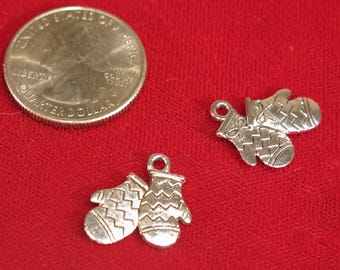 May include: Two silver-colored mitten charms with intricate detailing, next to a quarter coin, on a red background. The charms are small, with a loop at the top for attaching to jewelry or other crafts. The mittens have a textured design.