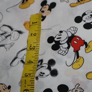 May include: White fabric featuring a repeating pattern of Mickey Mouse in various poses. Mickey is depicted in black, white, red, and yellow. A yellow measuring tape is placed on the fabric, showing measurements in inches.
