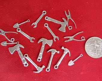 May include: A collection of silver-toned metal charms shaped like various tools, including hammers, axes, shovels, wrenches, and pliers. The charms are arranged on a red surface.