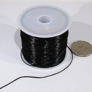 May include: A spool of black elastic cord wound around a white plastic bobbin. The cord is about 1/4 inch thick and is used for jewelry making or other crafts.